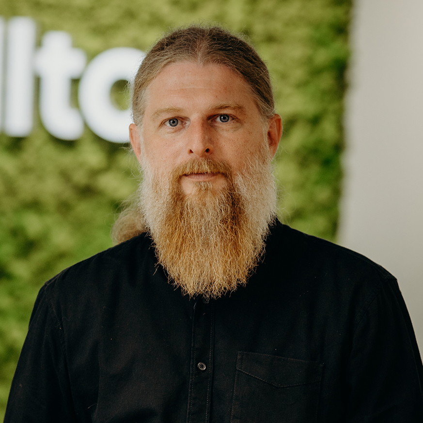 A man with a long beard standing in the office in front of the Kellton sign, wearing a black shirt.