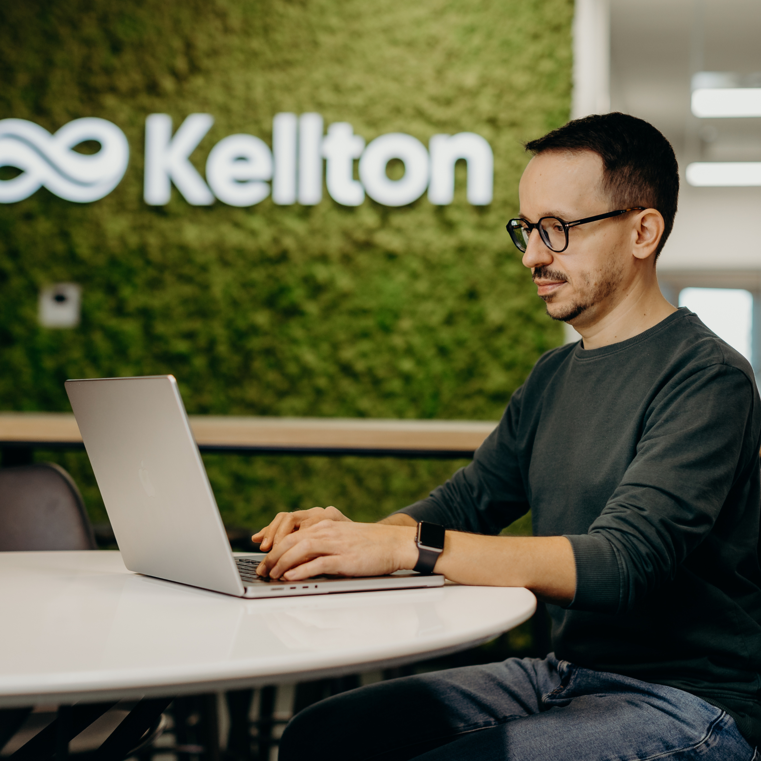 A man sitting in the office in front of the Kellton sign working on a laptop.